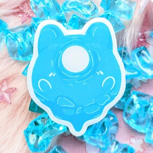 Puede incluir: Una pegatina azul y blanca de un personaje de dibujos animados con un ojo redondo y blanco. La pegatina está sobre un fondo rosa y blanco con gemas de plástico azul.