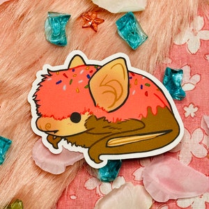 Könnte beinhalten: Ein rosa-brauner Fledermaus-Aufkleber mit rosa Zuckerguss und bunten Streuseln. Der Aufkleber befindet sich auf einem rosa-weißen Hintergrund.