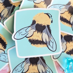 Könnte beinhalten: Ein Aufkleber mit einem Cartoon-Hummel mit schwarzen und gelben Streifen, blauen Flügeln und einem flauschigen Körper. Die Hummel befindet sich auf einem hellblauen Hintergrund.