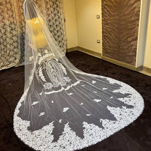 Puede incluir: Un velo de novia blanco largo con intrincados detalles de encaje. El velo presenta un borde de encaje floral detallado y una imagen bordada central. El velo se exhibe en un maniquí, mostrando su longitud y diseño.