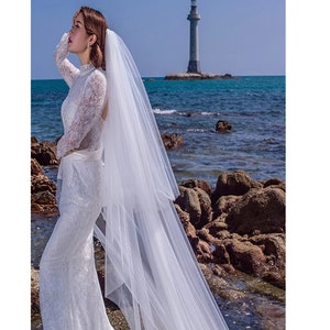 Puede incluir: Una novia con un vestido de novia de encaje blanco con un velo largo, de pie en una playa rocosa con un faro en el fondo.