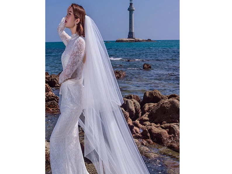 Puede incluir: Una novia con un vestido de novia de encaje blanco con un velo largo, de pie en una playa rocosa con un faro en el fondo.