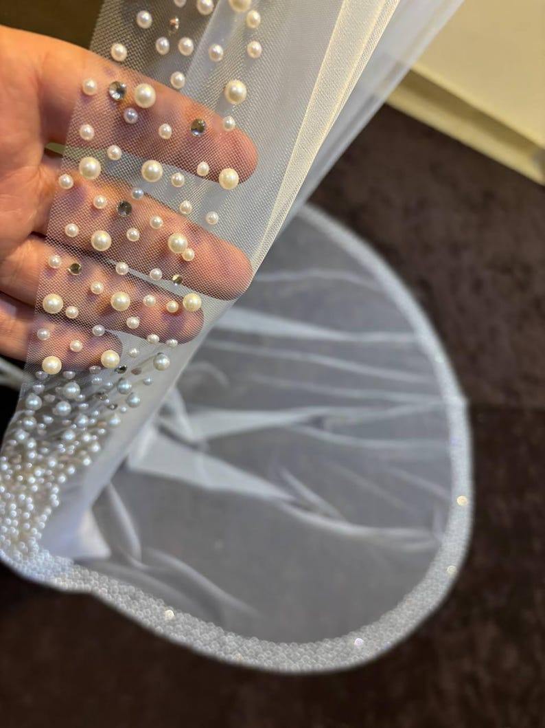 Peut inclure: Un voile de mari&eacute;e blanc orn&eacute; de fausses perles et de cristaux. Le bord du voile est &eacute;galement orn&eacute; de perles, cr&eacute;ant un design d&eacute;licat et &eacute;l&eacute;gant. Le voile est fait d'un tissu transparent et l&eacute;ger.