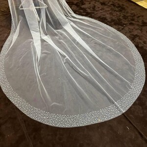 Peut inclure: Un voile de mari&eacute;e blanc transparent avec un bord arrondi. Le bord est orn&eacute; de petites perles scintillantes espac&eacute;es r&eacute;guli&egrave;rement. Le voile est drap&eacute; sur une surface marron fonc&eacute;, mettant en valeur sa texture d&eacute;licate.