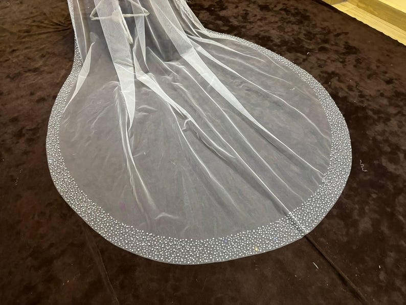 Peut inclure: Un voile de mari&eacute;e blanc transparent avec un bord arrondi. Le bord est orn&eacute; de petites perles scintillantes espac&eacute;es r&eacute;guli&egrave;rement. Le voile est drap&eacute; sur une surface marron fonc&eacute;, mettant en valeur sa texture d&eacute;licate.