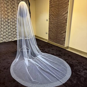 Peut inclure: Un long voile de mari&eacute;e blanc avec un bord perl&eacute;. Le voile est drap&eacute; sur un mannequin, mettant en valeur sa longueur et son design d&eacute;licat. La bordure perl&eacute;e ajoute une touche d'&eacute;l&eacute;gance au tissu transparent.