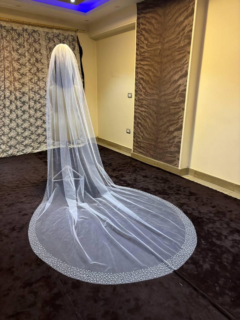 Peut inclure: Un long voile de mari&eacute;e blanc avec un bord perl&eacute;. Le voile est drap&eacute; sur un mannequin, mettant en valeur sa longueur et son design d&eacute;licat. La bordure perl&eacute;e ajoute une touche d'&eacute;l&eacute;gance au tissu transparent.