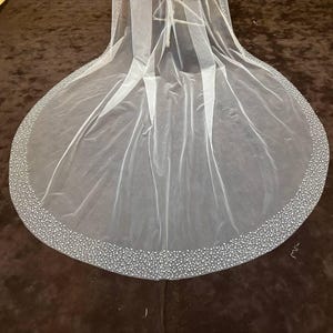 Peut inclure: Un voile de mari&eacute;e blanc transparent avec une bordure perl&eacute;e. Le voile est long et fluide, avec un bord arrondi. La bordure est dens&eacute;ment d&eacute;cor&eacute;e de petites perles rondes, ajoutant une touche d'&eacute;clat et d'&eacute;l&eacute;gance.