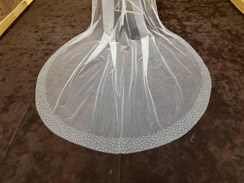 Peut inclure: Un voile de mari&eacute;e blanc transparent avec une bordure perl&eacute;e. Le voile est long et fluide, avec un bord arrondi. La bordure est dens&eacute;ment d&eacute;cor&eacute;e de petites perles rondes, ajoutant une touche d'&eacute;clat et d'&eacute;l&eacute;gance.
