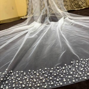 Peut inclure: Un voile de mari&eacute;e blanc transparent, orn&eacute; d'une bordure de perles et de petits cristaux. Le voile est fait d'un tissu fin et translucide. Les perles sont de diff&eacute;rentes tailles, cr&eacute;ant un effet textur&eacute;. Id&eacute;al pour un mariage.