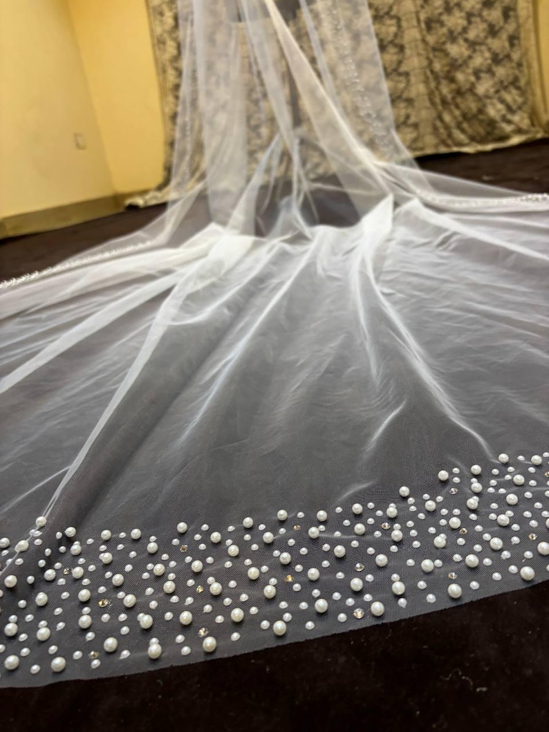 Peut inclure: Un voile de mari&eacute;e blanc transparent, orn&eacute; d'une bordure de perles et de petits cristaux. Le voile est fait d'un tissu fin et translucide. Les perles sont de diff&eacute;rentes tailles, cr&eacute;ant un effet textur&eacute;. Id&eacute;al pour un mariage.