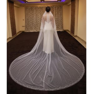 Peut inclure: Une robe de mariée blanche avec un long voile flottant. Le voile est orné de petits embellissements scintillants sur les bords. La robe présente un design simple et élégant, idéal pour une cérémonie de mariage.