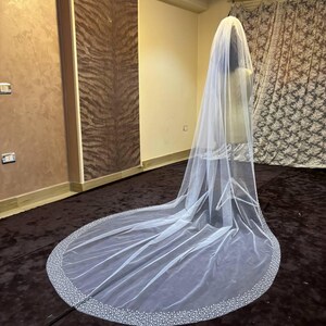 Peut inclure: Un long voile de mari&eacute;e blanc avec un bord perl&eacute;. Le voile est fait d'un tissu transparent et descend jusqu'au sol. Le bord est orn&eacute; de petites perles espac&eacute;es, cr&eacute;ant une bordure scintillante. Le voile est pr&eacute;sent&eacute; sur un mannequin.