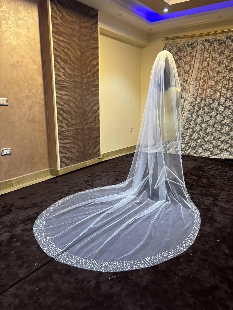 Peut inclure: Un long voile de mari&eacute;e blanc avec un bord perl&eacute;. Le voile est fait d'un tissu transparent et descend jusqu'au sol. Le bord est orn&eacute; de petites perles espac&eacute;es, cr&eacute;ant une bordure scintillante. Le voile est pr&eacute;sent&eacute; sur un mannequin.