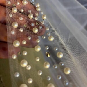 Peut inclure: Gros plan sur un tissu transparent blanc cass&eacute; orn&eacute; de fausses perles de diff&eacute;rentes tailles et de strass iris&eacute;s &eacute;pars. Les perles sont de couleur cr&egrave;me et ajoutent une touche d'&eacute;l&eacute;gance au mat&eacute;riau d&eacute;licat. Le tissu est probablement utilis&eacute; pour les v&ecirc;tements de mari&eacute;e.