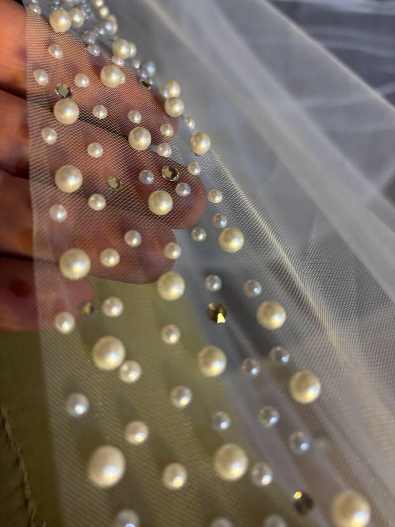 Peut inclure: Gros plan sur un tissu transparent blanc cass&eacute; orn&eacute; de fausses perles de diff&eacute;rentes tailles et de strass iris&eacute;s &eacute;pars. Les perles sont de couleur cr&egrave;me et ajoutent une touche d'&eacute;l&eacute;gance au mat&eacute;riau d&eacute;licat. Le tissu est probablement utilis&eacute; pour les v&ecirc;tements de mari&eacute;e.