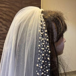 Peut inclure: Un voile de mari&eacute;e en tulle blanc orn&eacute; de perles et de cristaux. Le voile est fix&eacute; &agrave; un peigne et retombe gracieusement. Les perles sont de tailles vari&eacute;es, ajoutant &agrave; l'&eacute;l&eacute;gance du voile. Un accessoire de mariage classique.
