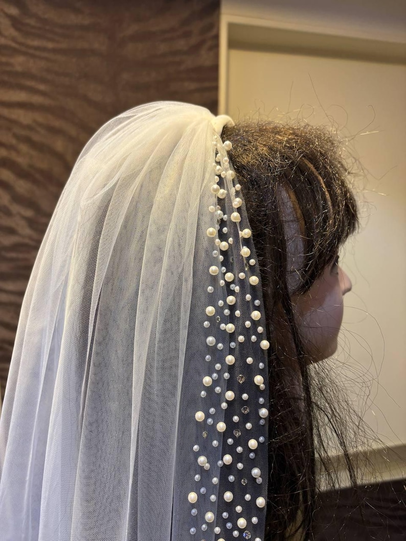Peut inclure: Un voile de mari&eacute;e en tulle blanc orn&eacute; de perles et de cristaux. Le voile est fix&eacute; &agrave; un peigne et retombe gracieusement. Les perles sont de tailles vari&eacute;es, ajoutant &agrave; l'&eacute;l&eacute;gance du voile. Un accessoire de mariage classique.