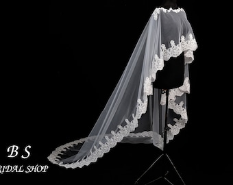 May include: A white bridal veil with a delicate lace trim. The veil is displayed on a black mannequin against a black background. The text "BS BRIDAL SHOP" is in the lower left corner.