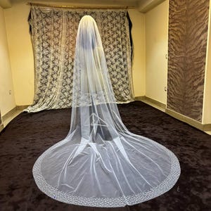 Peut inclure: Un long voile de mari&eacute;e blanc et fluide avec un bord perl&eacute;. Le voile est fait d'un tissu transparent et descend jusqu'au sol. Le bord est orn&eacute; de petites perles scintillantes. Le voile est pr&eacute;sent&eacute; dans une pi&egrave;ce avec une moquette marron et des rideaux &agrave; motifs.