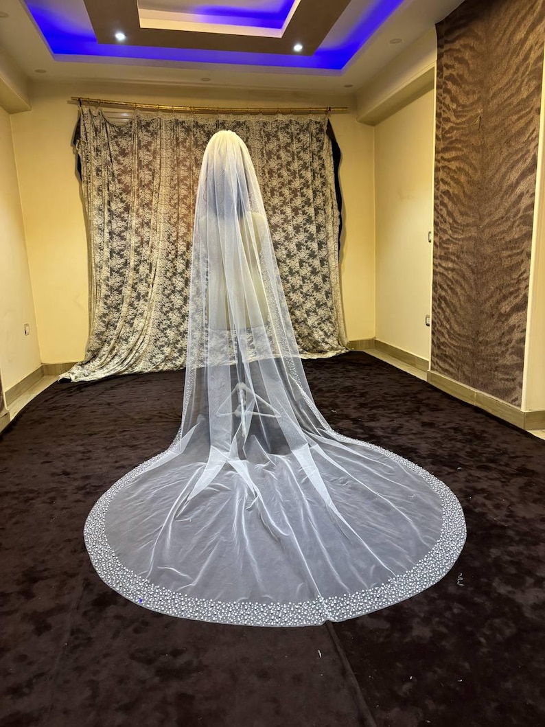 Peut inclure: Un long voile de mari&eacute;e blanc et fluide avec un bord perl&eacute;. Le voile est fait d'un tissu transparent et descend jusqu'au sol. Le bord est orn&eacute; de petites perles scintillantes. Le voile est pr&eacute;sent&eacute; dans une pi&egrave;ce avec une moquette marron et des rideaux &agrave; motifs.