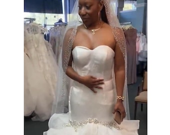May include: A bride wearing a white strapless wedding dress with a beaded veil and a ruffled skirt. The dress has a fitted bodice and a mermaid silhouette.