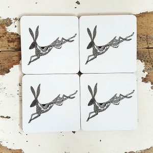 May include: Set of four white square coasters with a black and white illustration of a stylized hare leaping. The hare is detailed with a patterned design.