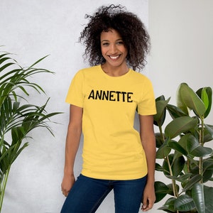 Puede incluir: Una mujer lleva una camiseta amarilla de manga corta con la palabra "ANNETTE" impresa en negro en la parte delantera. Est&aacute; de pie frente a una pared blanca con plantas a cada lado.