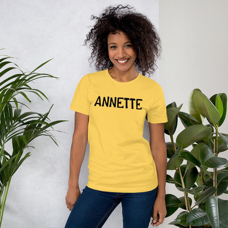 Puede incluir: Una mujer lleva una camiseta amarilla de manga corta con la palabra "ANNETTE" impresa en negro en la parte delantera. Est&aacute; de pie frente a una pared blanca con plantas a cada lado.