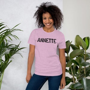 Puede incluir: Una mujer lleva una camiseta rosa con la palabra "ANNETTE" impresa en negro en la parte delantera. Est&aacute; de pie frente a una pared blanca con plantas a cada lado.