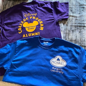 May include: Two t-shirts, one purple with yellow text that reads "Disney College Program Spring 2010 Alumni" and the other blue with white text that reads "Michael Rutgers University" and a white castle graphic.