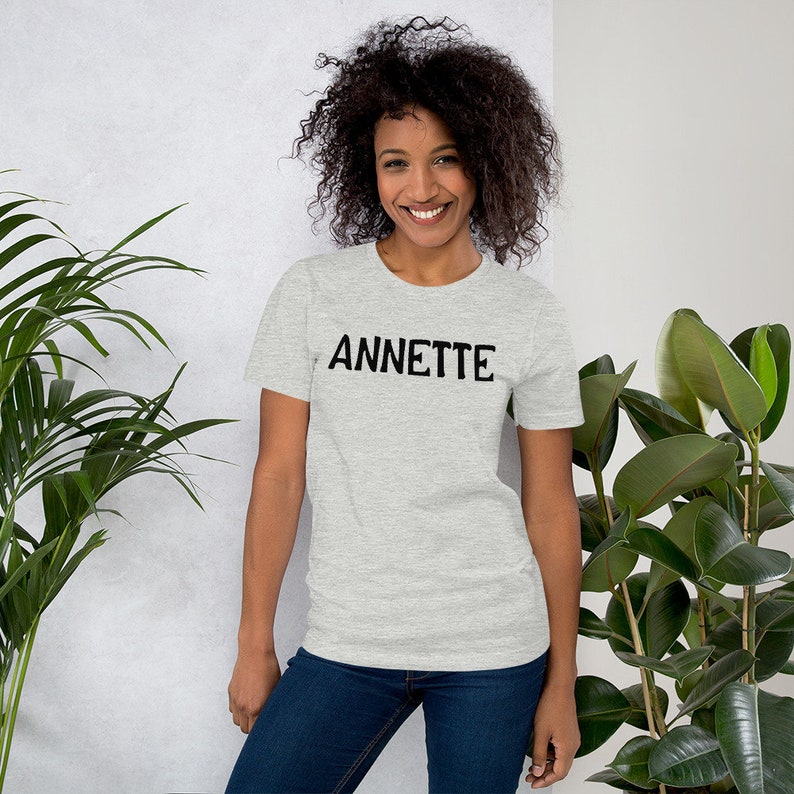 Puede incluir: Una mujer lleva una camiseta gris jaspeada con la palabra "ANNETTE" impresa en negro en la parte delantera. Est&aacute; sonriendo y mirando a la c&aacute;mara.