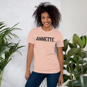 Puede incluir: Una mujer con una camiseta rosa claro con la palabra "ANNETTE" impresa en negro en la parte delantera. Lleva pantalones vaqueros azules y tiene el pelo rizado casta&ntilde;o.