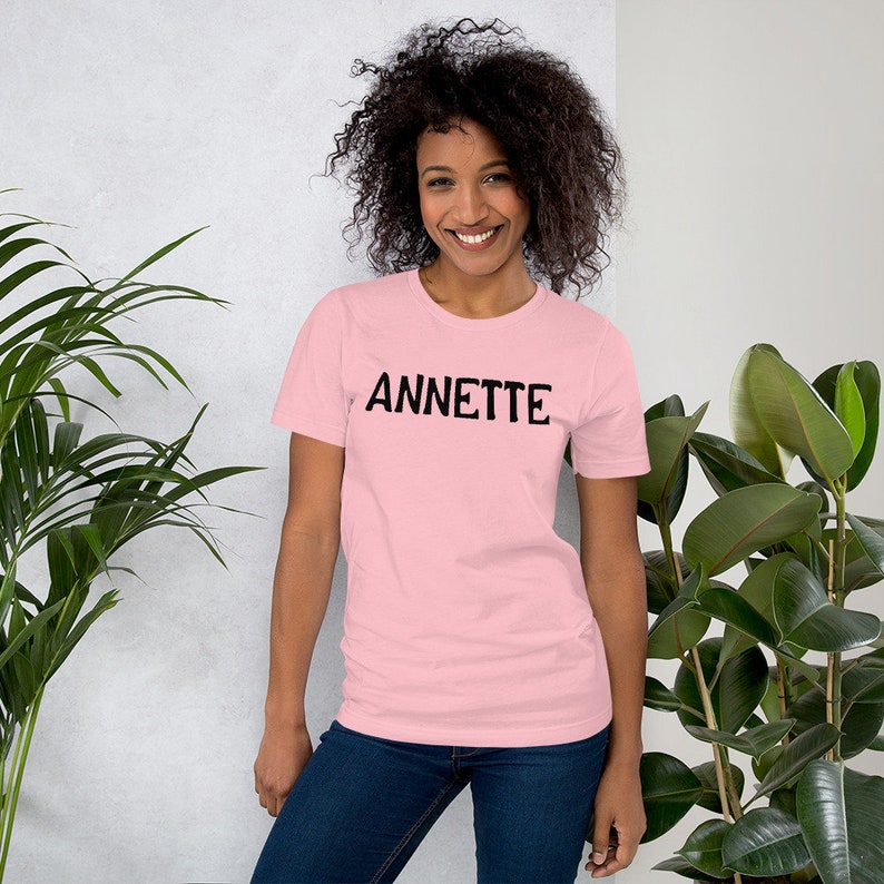 Puede incluir: Una camiseta rosa de manga corta con la palabra "ANNETTE" impresa en negro en la parte delantera.