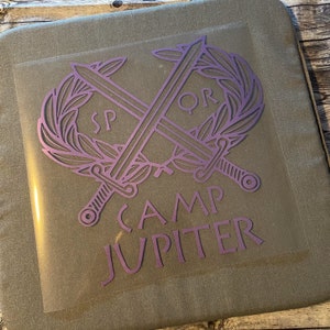 May include: A purple heat transfer vinyl design featuring crossed swords, a laurel wreath, and the text "SPQR" and "CAMP JUPITER". The design is on a clear carrier sheet, ready for application. The image is on a neutral background.