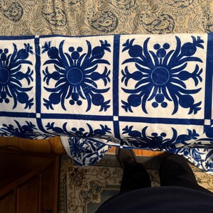 Puede incluir: Una manta de tiro blanca y azul con un dise&ntilde;o repetido de pi&ntilde;as estilizadas. La manta est&aacute; doblada y drapeada sobre una cama.