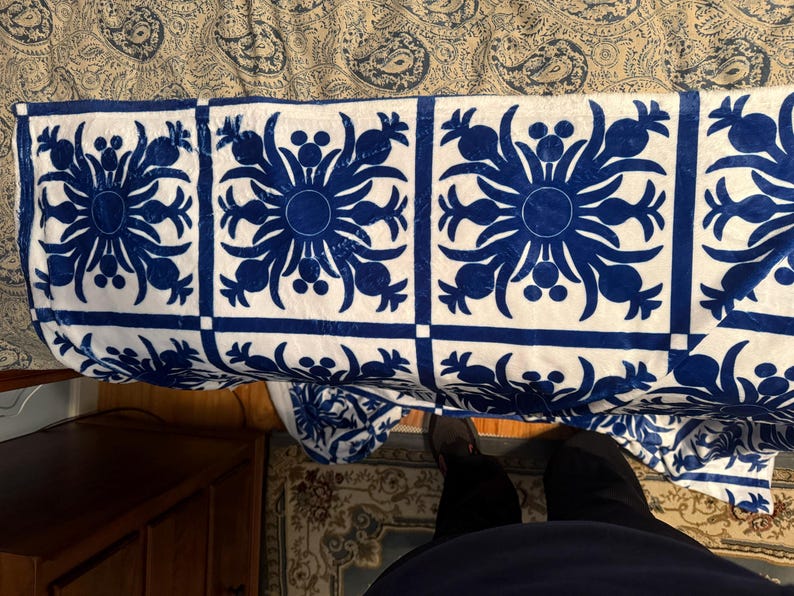 Puede incluir: Una manta de tiro blanca y azul con un dise&ntilde;o repetido de pi&ntilde;as estilizadas. La manta est&aacute; doblada y drapeada sobre una cama.