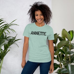 Puede incluir: Una mujer con una camiseta azul claro con la palabra "ANNETTE" impresa en negro en la parte delantera. Ella sonr&iacute;e y mira a la c&aacute;mara.