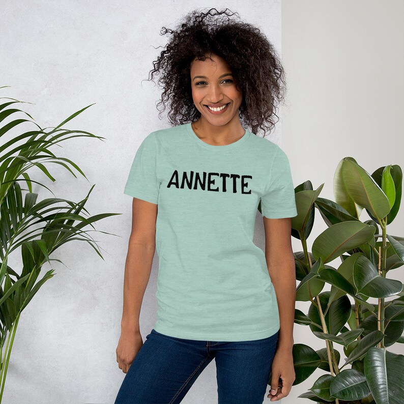 Puede incluir: Una mujer con una camiseta azul claro con la palabra "ANNETTE" impresa en negro en la parte delantera. Ella sonr&iacute;e y mira a la c&aacute;mara.