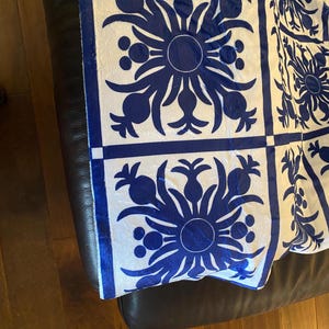Puede incluir: Una tela con estampado blanco y azul con un dise&ntilde;o repetido de pi&ntilde;as y flores. La tela est&aacute; doblada y drapeada sobre una superficie de cuero marr&oacute;n.
