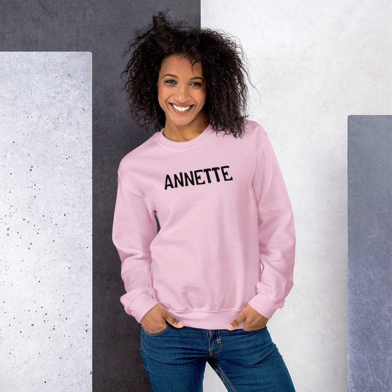 Op de afbeelding: Een roze sweatshirt met ronde hals en de naam "ANNETTE" in het zwart op de voorkant.