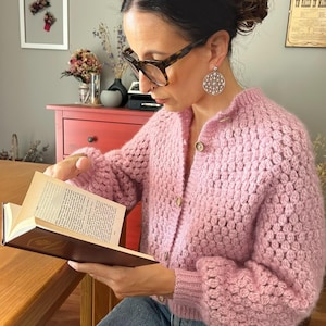 Könnte beinhalten: Eine hellrosa, langärmelige Strickjacke mit Knopfleiste, die durchgehend mit einem strukturierten Noppenmuster versehen ist. Das Kleidungsstück hat einen Rundhalsausschnitt, gerippte Bündchen und helle Knöpfe auf der Vorderseite, getragen über einer blauen Jeans.
