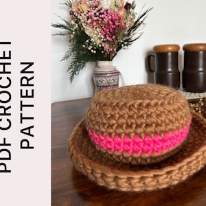 Può includere: Un cappello a uncinetto marrone con una fascia a uncinetto rosa. Il cappello è su un tavolo di legno.