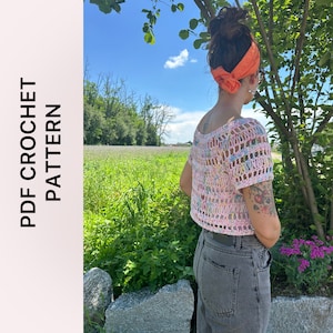 Può includere: Un top in crochet rosa e bianco con un motivo floreale colorato. Il top ha maniche corte e una lunghezza crop. La modella indossa il top con una bandana arancione brillante legata intorno alla testa.