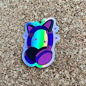 K&ouml;nnte beinhalten: Ein holografischer Aufkleber mit lila Kopfh&ouml;rern mit Katzenohren. Die Kopfh&ouml;rer haben einen regenbogenfarbenen, schillernden Hintergrund mit wei&szlig;en Sternen.