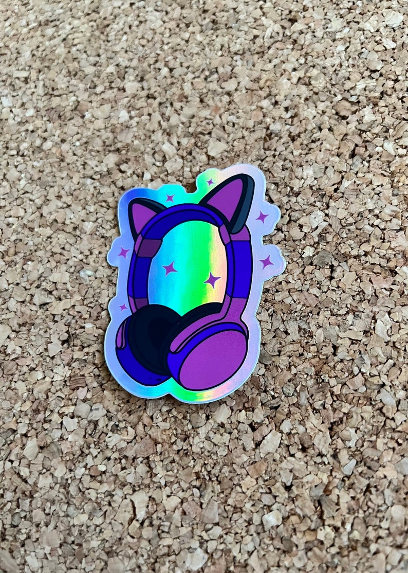 K&ouml;nnte beinhalten: Ein holografischer Aufkleber mit lila Kopfh&ouml;rern mit Katzenohren. Die Kopfh&ouml;rer haben einen regenbogenfarbenen, schillernden Hintergrund mit wei&szlig;en Sternen.