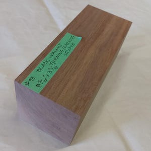 Peut inclure: Un bloc rectangulaire en noyer noir avec une étiquette verte indiquant "#93 BLACK WALNUT TURNING CARVING SQUARE 8 5/16 + 3 5/16". Le bois est brun rougeâtre avec une teinte violette à l'extrémité.