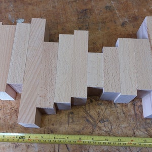 May include: A collection of 14 wooden blocks of varying sizes and widths, arranged in a staggered pattern on a wooden surface. The blocks are all made of light-coloured wood and have a smooth, polished finish.