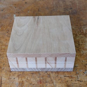 Op de afbeelding: Een lichtgekleurde houten doos met een vierkante vorm. De bovenkant is een massief stuk hout, terwijl de zijkanten een decoratief patroon van afwisselende stukken hout hebben. De doos staat op een houten oppervlak.