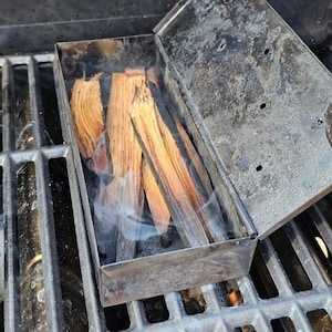 May include: A metal smoker box with wood chips inside, placed on a grill. The box is open, and smoke is rising from the wood. The grill grates are visible, with flames underneath.